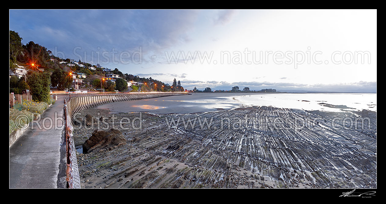 Image of Tahunanui Beach evening sunset panorama with dramatic geologial rock patterns in foreground, and Rocks Road, Moana, left, Nelson, Nelson City District, Nelson Region, New Zealand (NZ) stock photo image