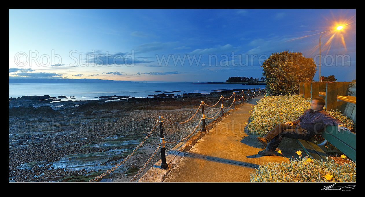 Image of Tahunanui Beach waterfront walkway along Rocks Road and Wakefield Quay, with person watching sunset over Tasman Bay. Panorama, Nelson, Nelson City District, Nelson Region, New Zealand (NZ) stock photo image