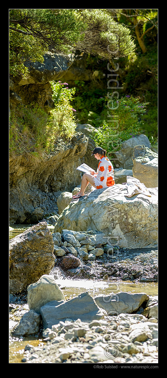 Image of Summer river swimming in Remutaka (Rimutaka) Forest Park. Woman sunbathing and reading a magazine on rocks after swim in freshwater pool. Sunny sunlit waterhole. Vertical panorama, Remutaka Forest Park, South Wairarapa District, Wellington Region, New Zealand (NZ) stock photo image