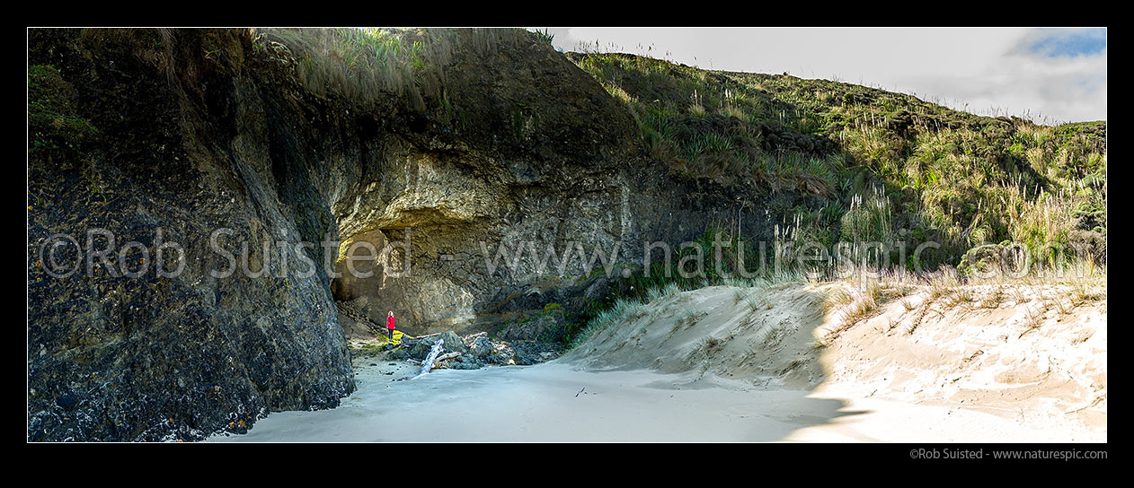 Image of Old sea cave on Mitimiti Beach, with visitor checking out the natural archway. Okaihau Pa and Moerewa Point. Panorama, Mitimiti, Far North District, Northland Region, New Zealand (NZ) stock photo image