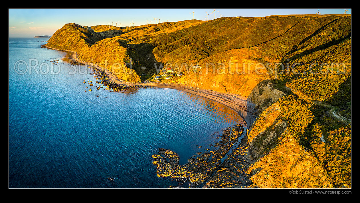 Image of Smiths Bay at Makara Beach, with baches nestled under the Mill Creek Wind farm turbines. Mana and Kapiti Islands above. Aerial panorama, Makara Beach, Wellington City District, Wellington Region, New Zealand (NZ) stock photo image