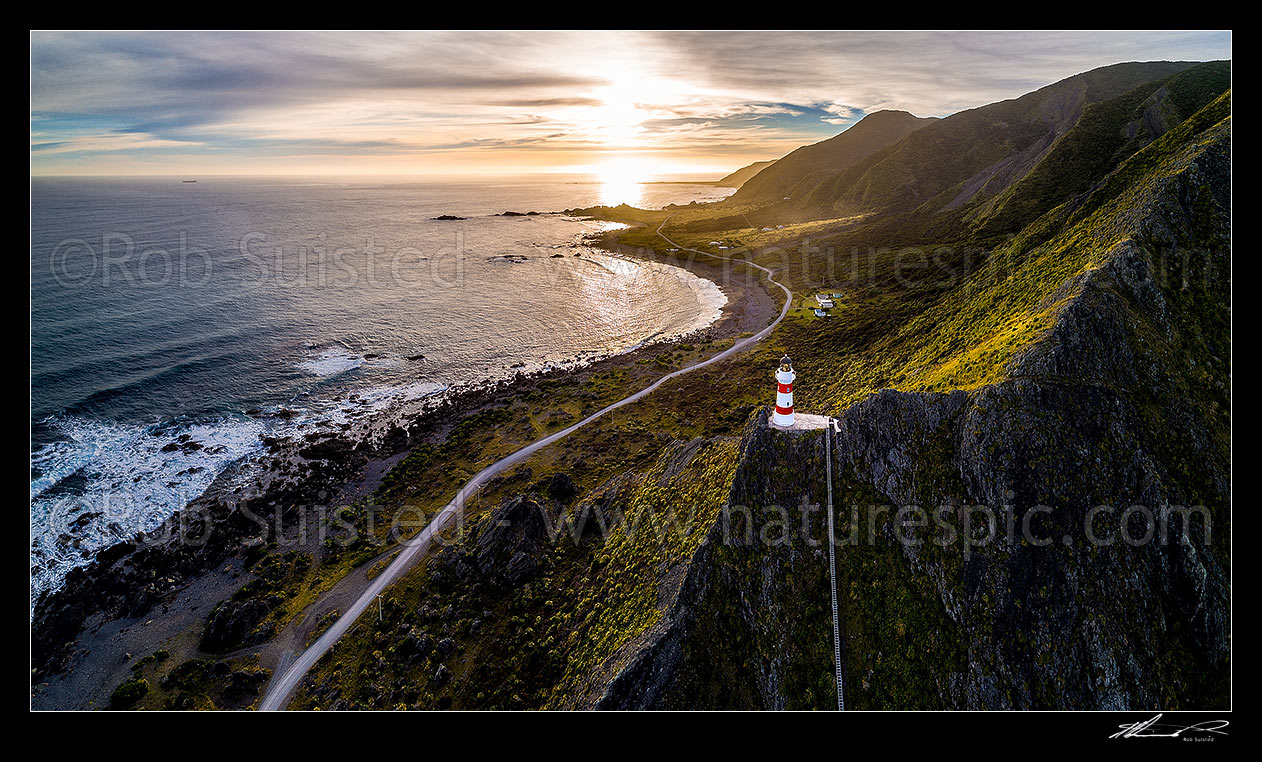 Image of Cape Palliser lighthouse above Palliser Bay baches and Kirikiri Bay on a moody sunset (Matakitakiakupe). Aerial panorama with golden evening sunset, Cape Palliser, South Wairarapa District, Wellington Region, New Zealand (NZ) stock photo image