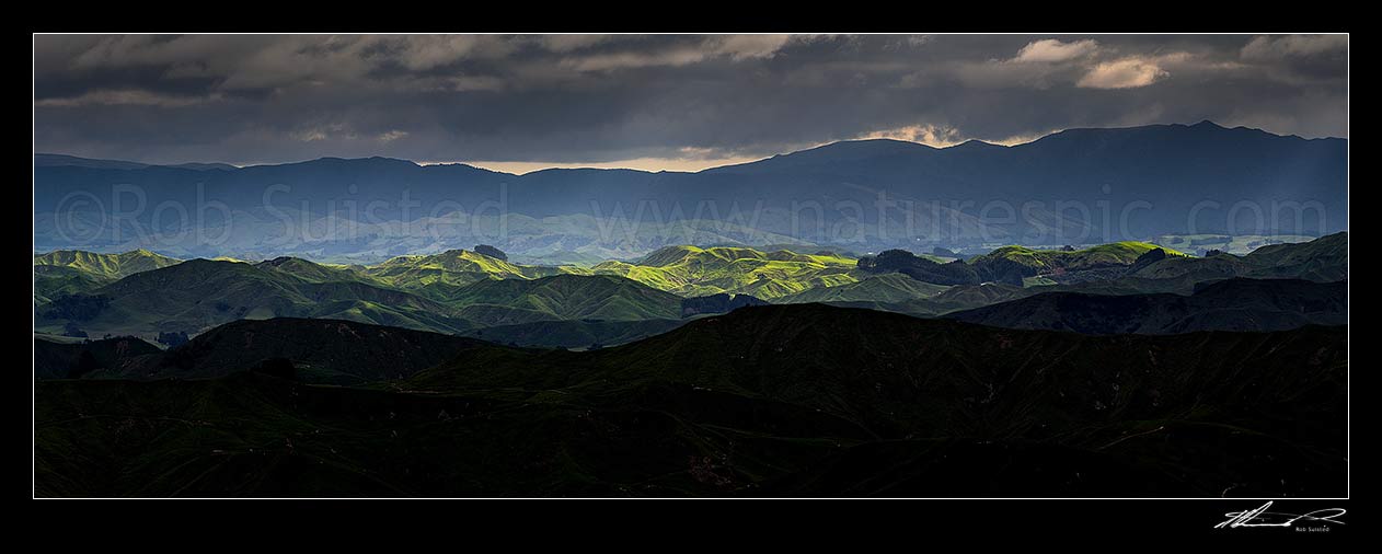 Image of Wairarapa hill country moody day, with breaking sunlight lighting hills and ridges near Eketahuna. Panorama view with northern Tararua Ranges behind. Northern Wairarapa, Pori, Tararua District, Manawatu-Wanganui Region, New Zealand (NZ) stock photo image