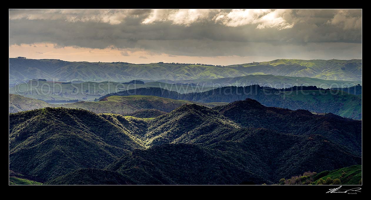 Image of Wairarapa hill country moody day, with breaking sunlight lighting hills and ridges near Eketahuna. Panorama view with northern Tararua Ranges behind. Northern Wairarapa, Pori, Tararua District, Manawatu-Wanganui Region, New Zealand (NZ) stock photo image