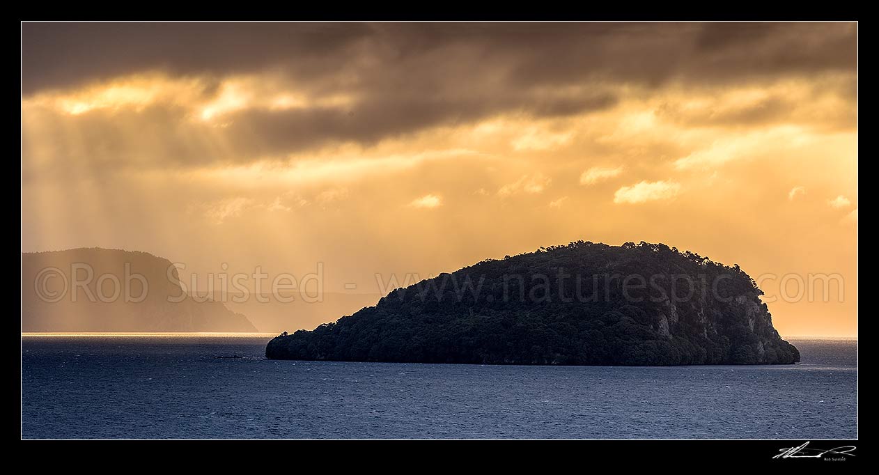 Image of Lake Taupo with moody weather breaking. Motutaiko Island in Lake Taupo (Taupomoana). Crepuscular rays on the western lake shore cliffs of Karangahape behind. Panorama, Motutere, Taupo District, Waikato Region, New Zealand (NZ) stock photo image